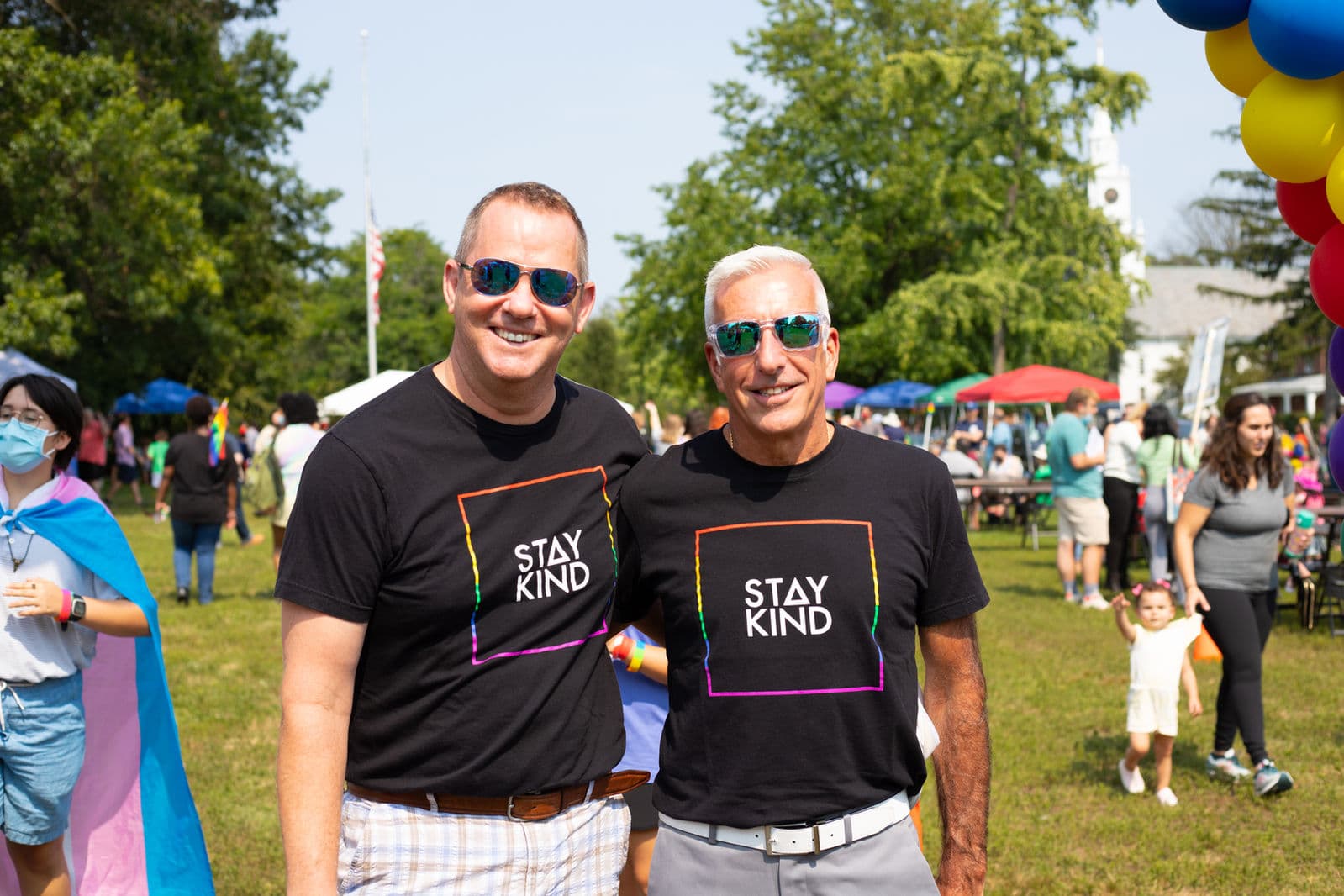 Two men in matching black 'Stay Kind' t-shirts stand arm in arm on the Town Green, with a rainbow balloon arch and the church steeple behind them