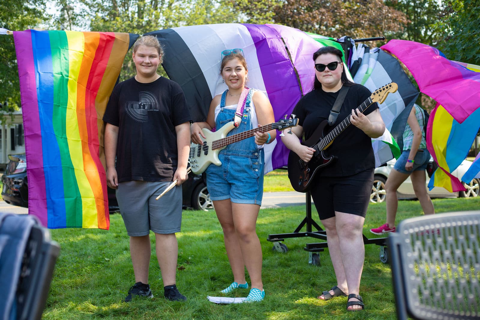 Three young musicians with a bass, an electric guitar and drumsticks stand in front of rainbow, asexual, genderqueer and pansexual flags strung up behind them