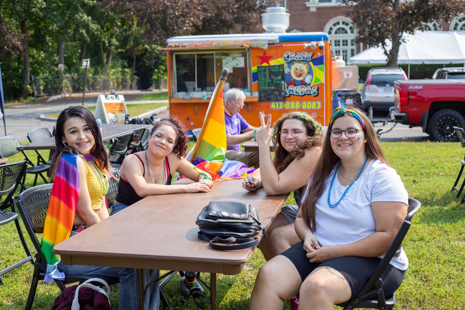 Four young festival-goers sit together at a folding table, one wrapped in a rainbow cape, with the Crazy Arepas food truck parked behind