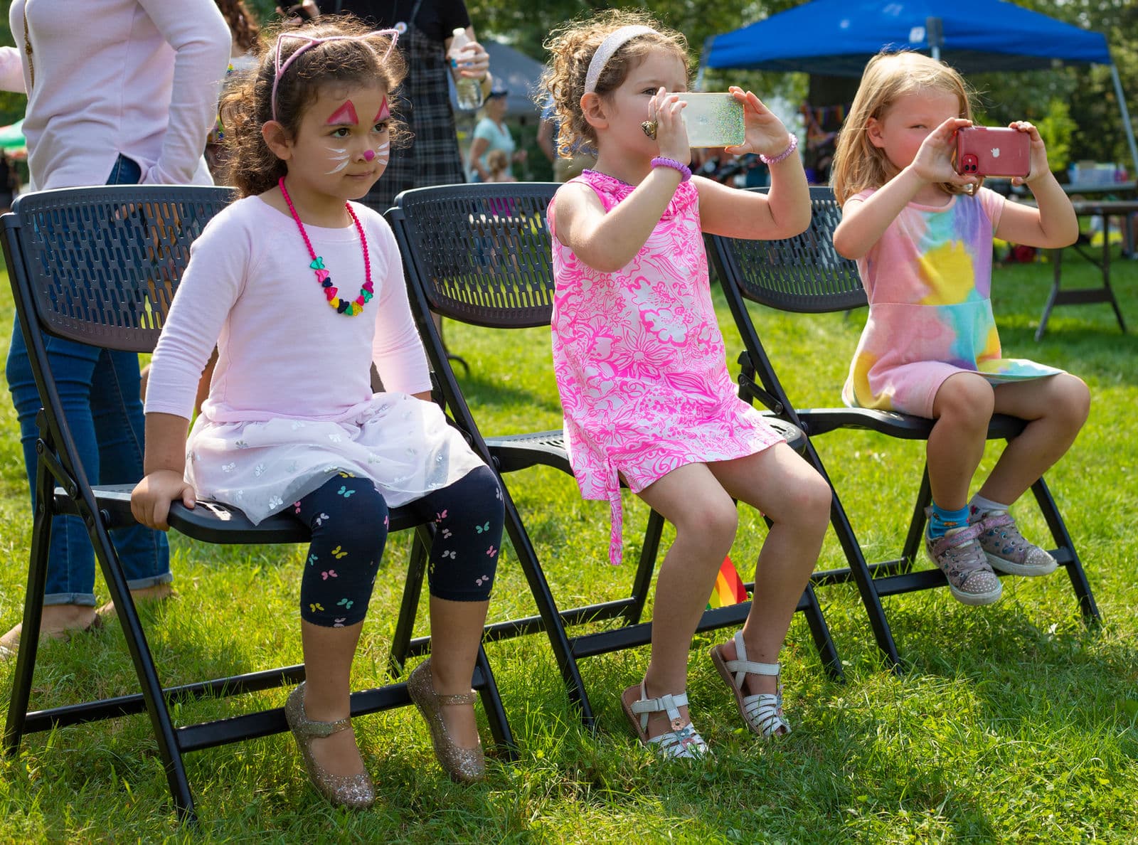 Three small children sit on folding chairs watching a performance; one has cat whiskers painted on their face and two hold up phones to take photos