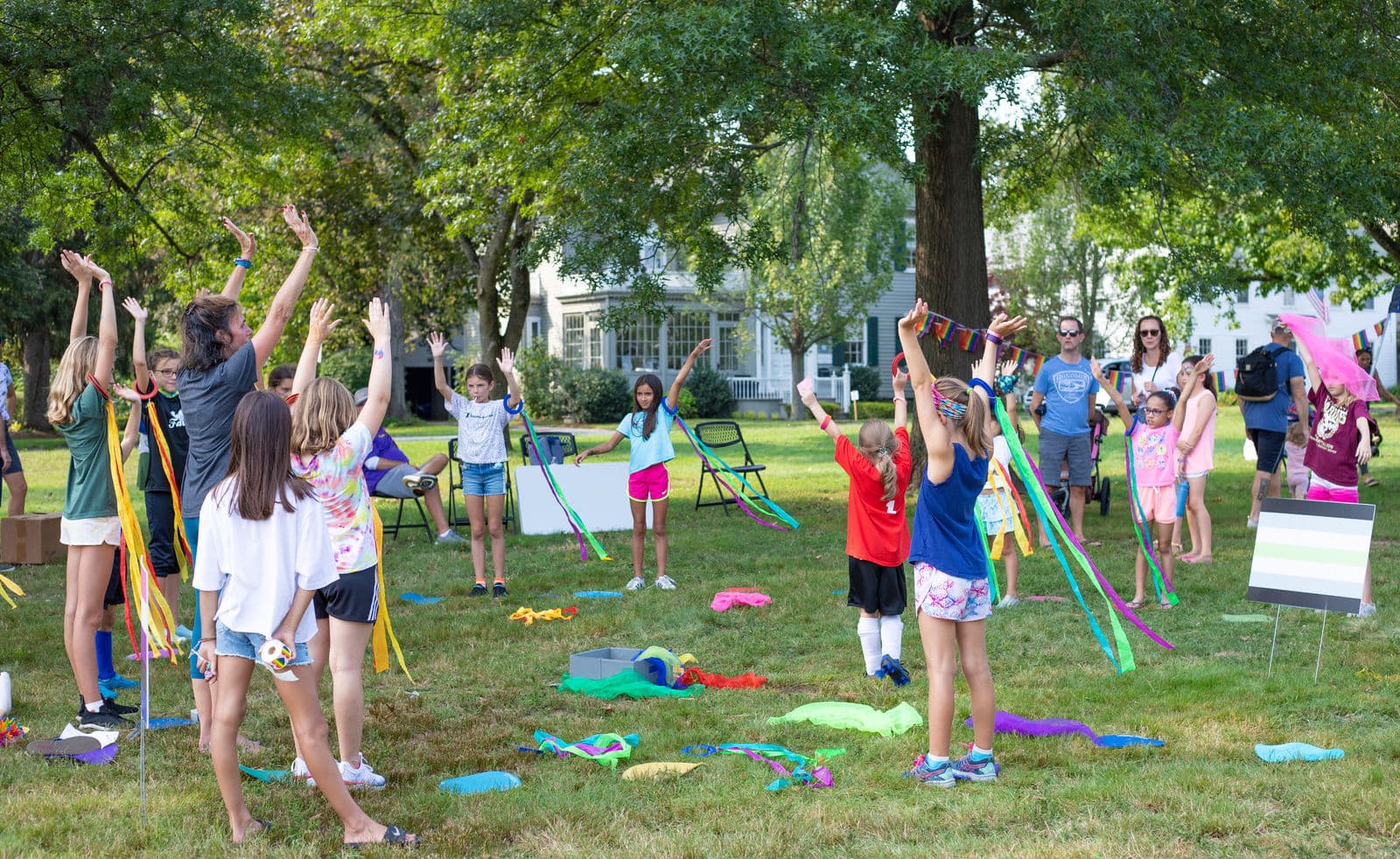 Children and adults raise colored ribbon streamers above their heads during a movement activity on the grass beneath the Green's trees