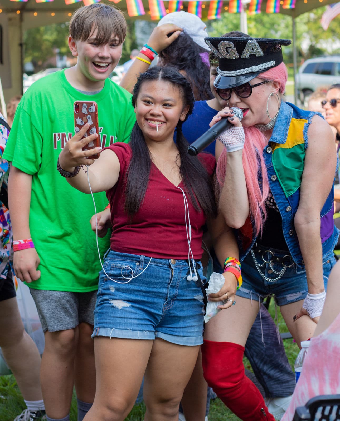 A festival-goer takes a selfie beside a performer with long pink hair singing into a microphone, wearing a denim vest paneled in rainbow colors