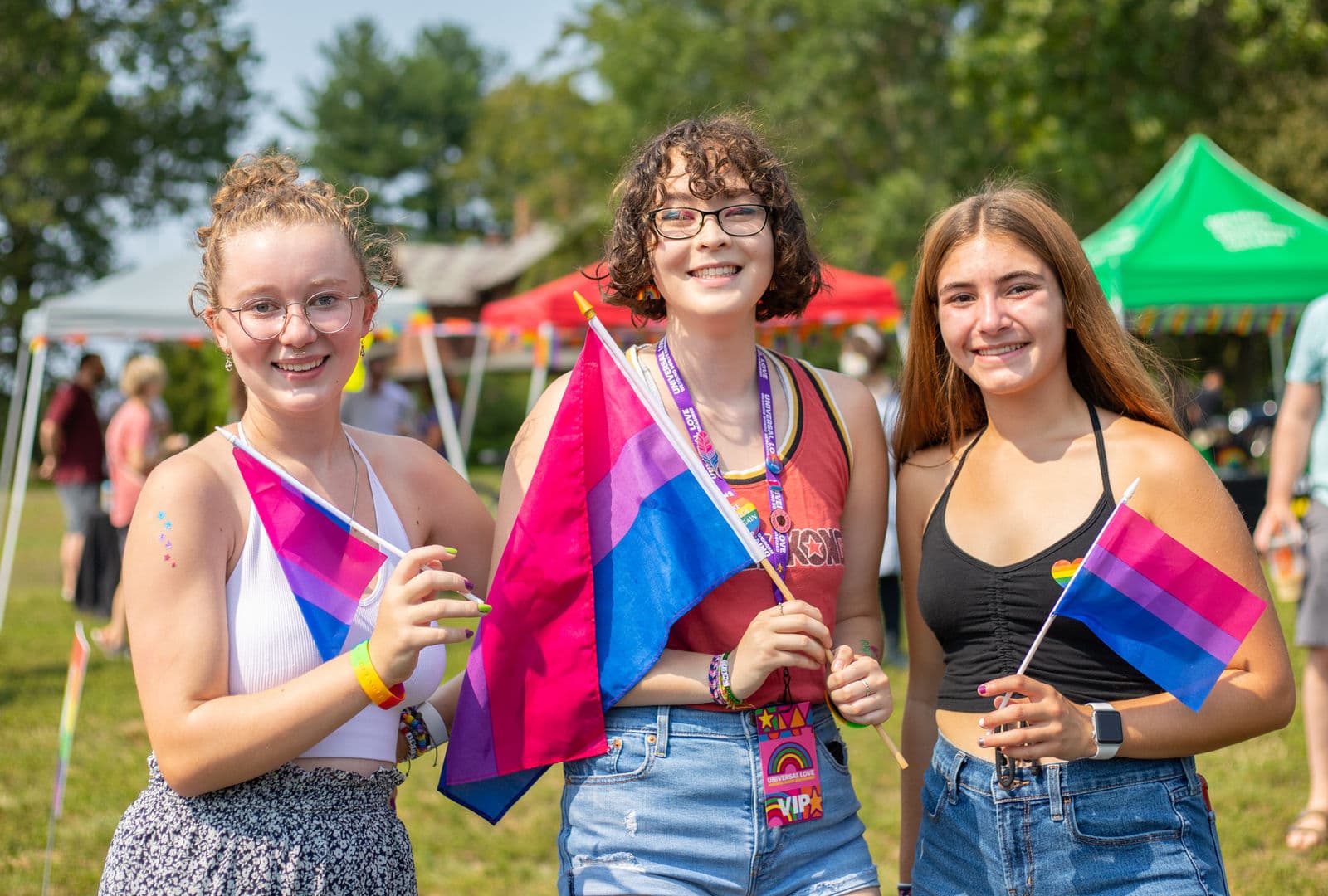 Three teenagers smile at the camera holding bisexual pride flags, with vendor tents in the background