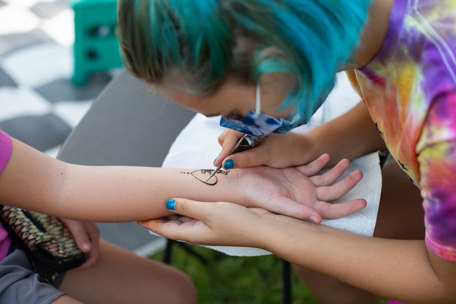 An artist with turquoise hair leans in to draw a henna heart on a visitor's forearm at a craft table