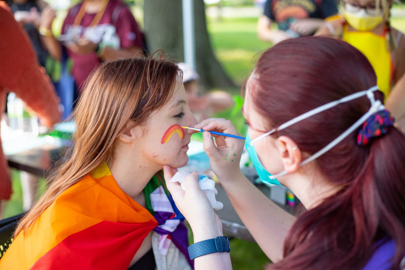 A volunteer paints a rainbow across the cheek of a visitor who is wearing a rainbow flag around their shoulders